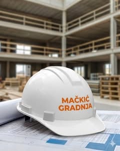 Bela građevinska kaciga sa natpisom „Mačkić Gradnja“ na gradilištu, sa nacrtima u prvom planu